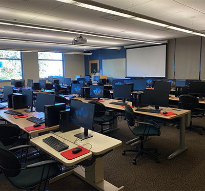 Chairs behind desks, each with a desktop computer and monitor, facing a screen and lecturn, windows outside are visible